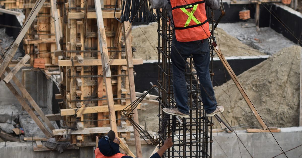 Two workers in safety vests climb a steel structure on a busy construction site.
